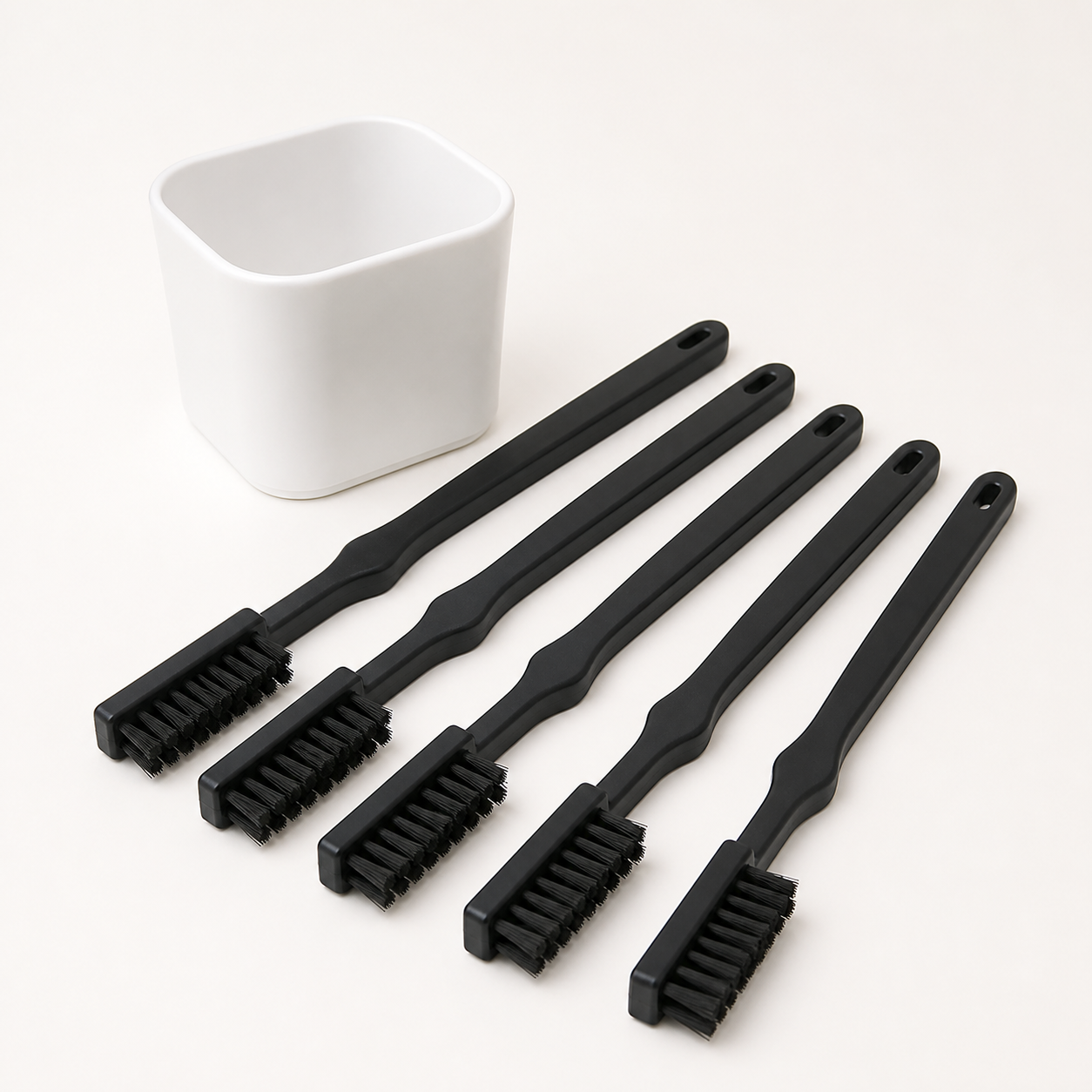 Five slim black hard-bristle crevice cleaning brushes arranged beside a white storage cup